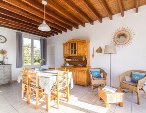 A dining area with a wooden table and chairs, a wooden cabinet, wicker chairs with cushions, and a sunburst wall decoration.