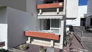 Ein kleiner Sitzbereich draußen mit Tisch und Stühlen, ein Fenster mit Vorhang und ein Schild an der Wand.