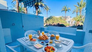 Ein Frühstückstisch mit Croissants, Obst und Getränken mit Blick auf einen Pool und Palmen.