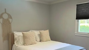 Ein ordentlich eingerichtetes Schlafzimmer mit einem Bett, einem Nachttisch, einer wandhängenden Makramee-Dekoration und einem Fenster mit Jalousie.