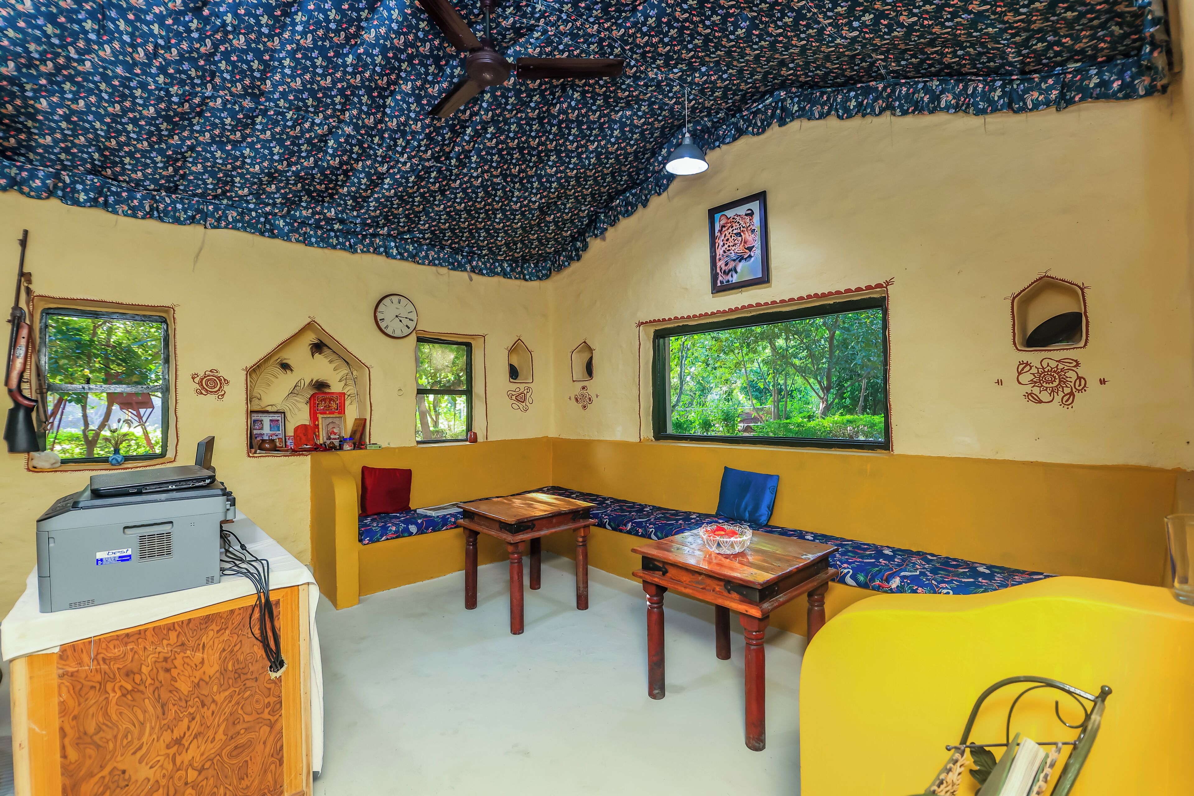 A room with a ceiling fan, a table with a bowl, a bench, and a window with a view of greenery.