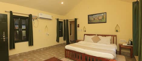 A hotel room with a bed, a wooden table, a small bedside table, a wall-mounted air conditioner, a window with curtains, and a painting on the wall.
