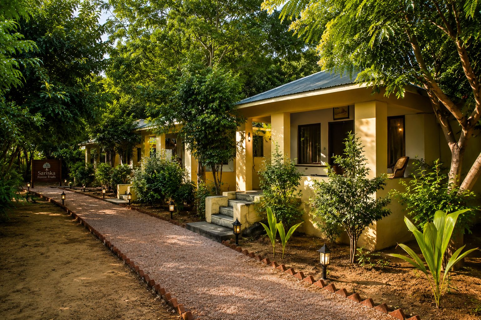 A row of yellow buildings with a gravel path leading to them, surrounded by trees and plants.