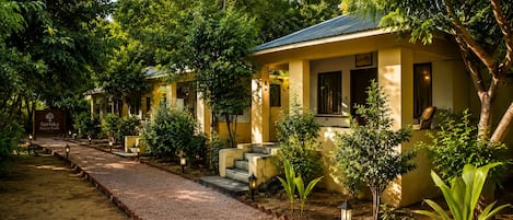 A row of yellow buildings with a gravel path leading to them, surrounded by trees and plants.