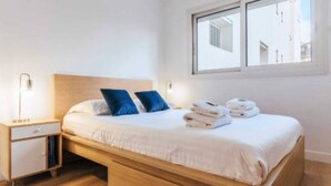 A single bed with a wooden headboard, white bedding, two blue pillows, a nightstand with a lamp, and a window with a view of buildings. - Cosy apartment: 2-minute walk from PSG & Roland-Garros (Boulogne-Billancourt)