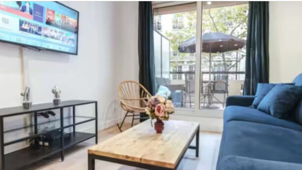 A living room with a blue sofa, a wooden coffee table, a TV on the wall, and a view of the outside balcony. - Cosy apartment: 2-minute walk from PSG & Roland-Garros (Boulogne-Billancourt)