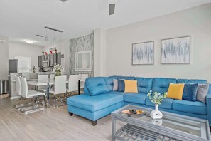 A modern living room with a blue sectional sofa, a dining area with white chairs, and a kitchen with grey cabinets.