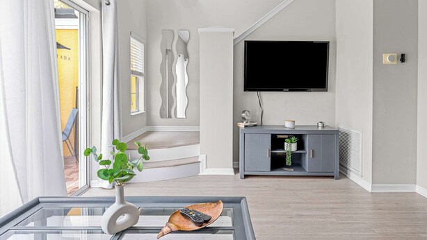 Een moderne woonkamer met een glazen salontafel, een flatscreen-televisie aan de muur bevestigd, een grijze entertainmentkast, en een trap naar een buitenruimte.