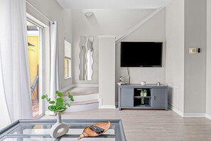 A modern living room with a glass coffee table, a flat-screen TV mounted on the wall, a grey entertainment unit, and a staircase leading to an outdoor area.