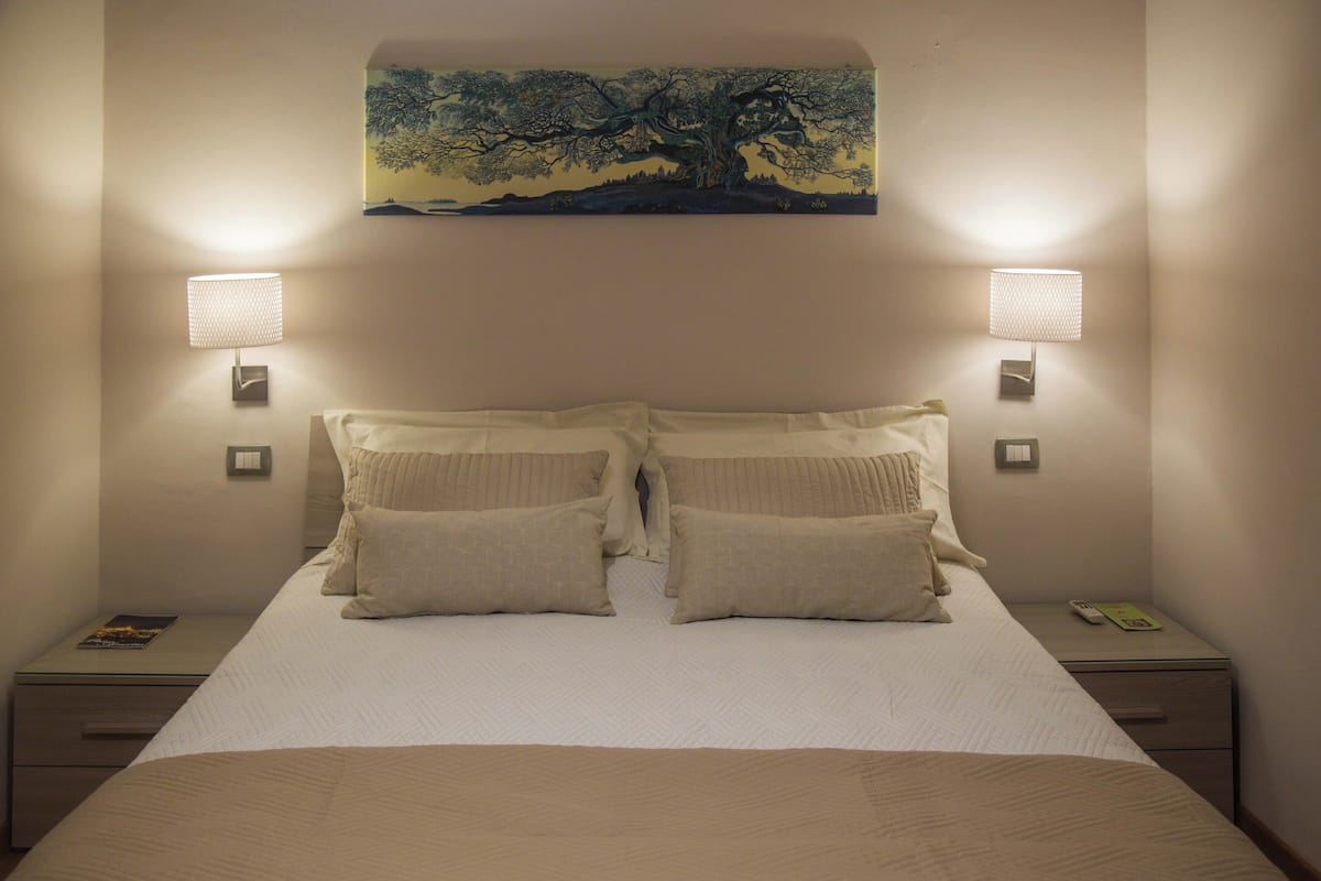 Una cama bien hecha con dos almohadas, un cuadro encima y dos lámparas de pared a cada lado.