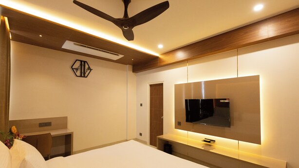 A modern hotel room with a large bed, a flat-screen TV mounted on the wall, a ceiling fan, and a bedside table with a lamp. - Kaizen Residency (Tiruchirappalli)