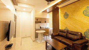 A modern living room with a brown leather sofa, a wooden coffee table, and a flat-screen TV. There is a ceiling fan, a kitchen area with a refrigerator, and a dining table with chairs. - Kaizen Residency (Tiruchirappalli)