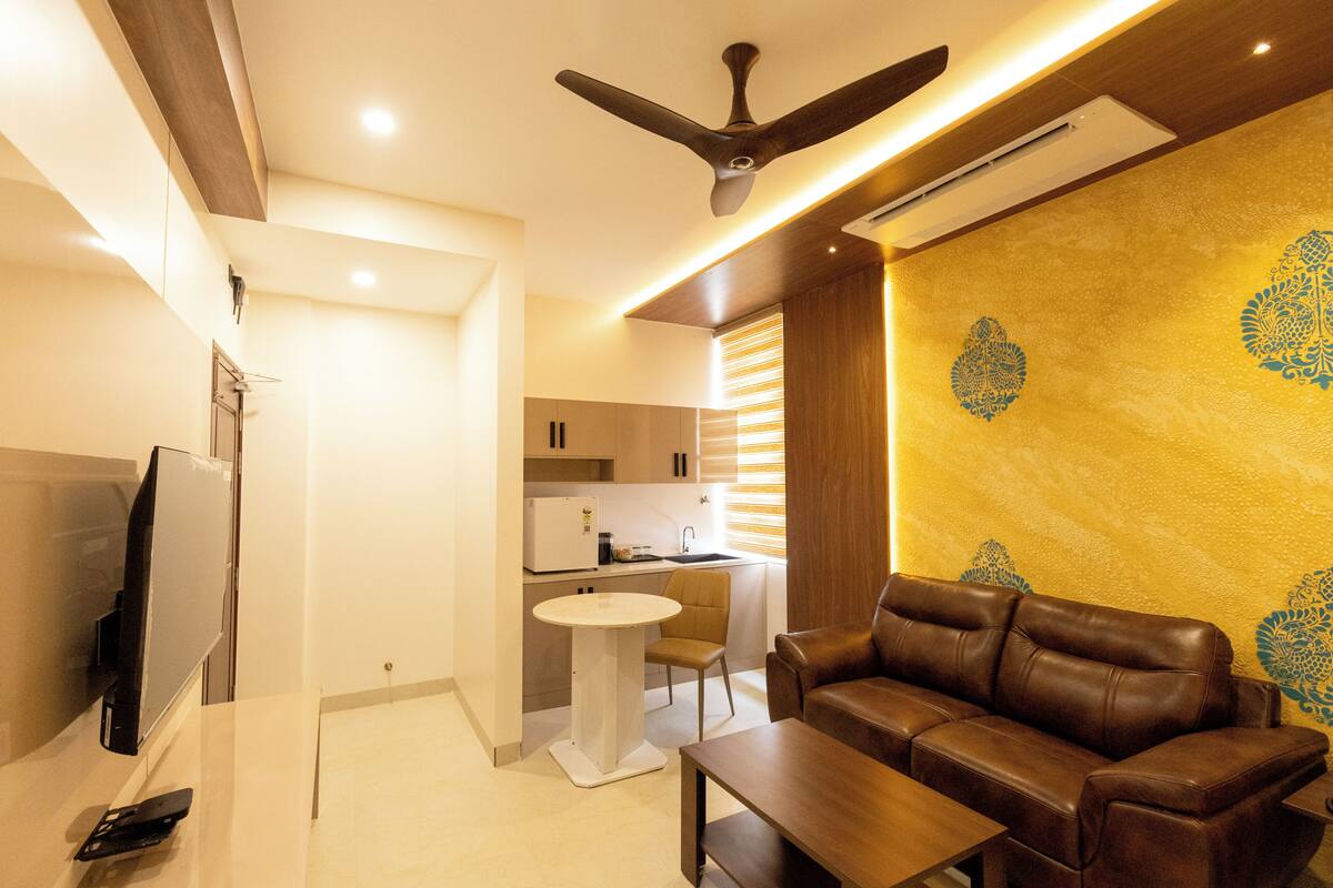 A modern living room with a brown leather sofa, a wooden coffee table, and a flat-screen TV. There is a ceiling fan, a kitchen area with a refrigerator, and a dining table with chairs.