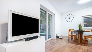 Ett modernt vardagsrum med en platt-TV, ett matbord med stolar, en väggklocka och en skjutdörr i glas som leder ut till en balkong.