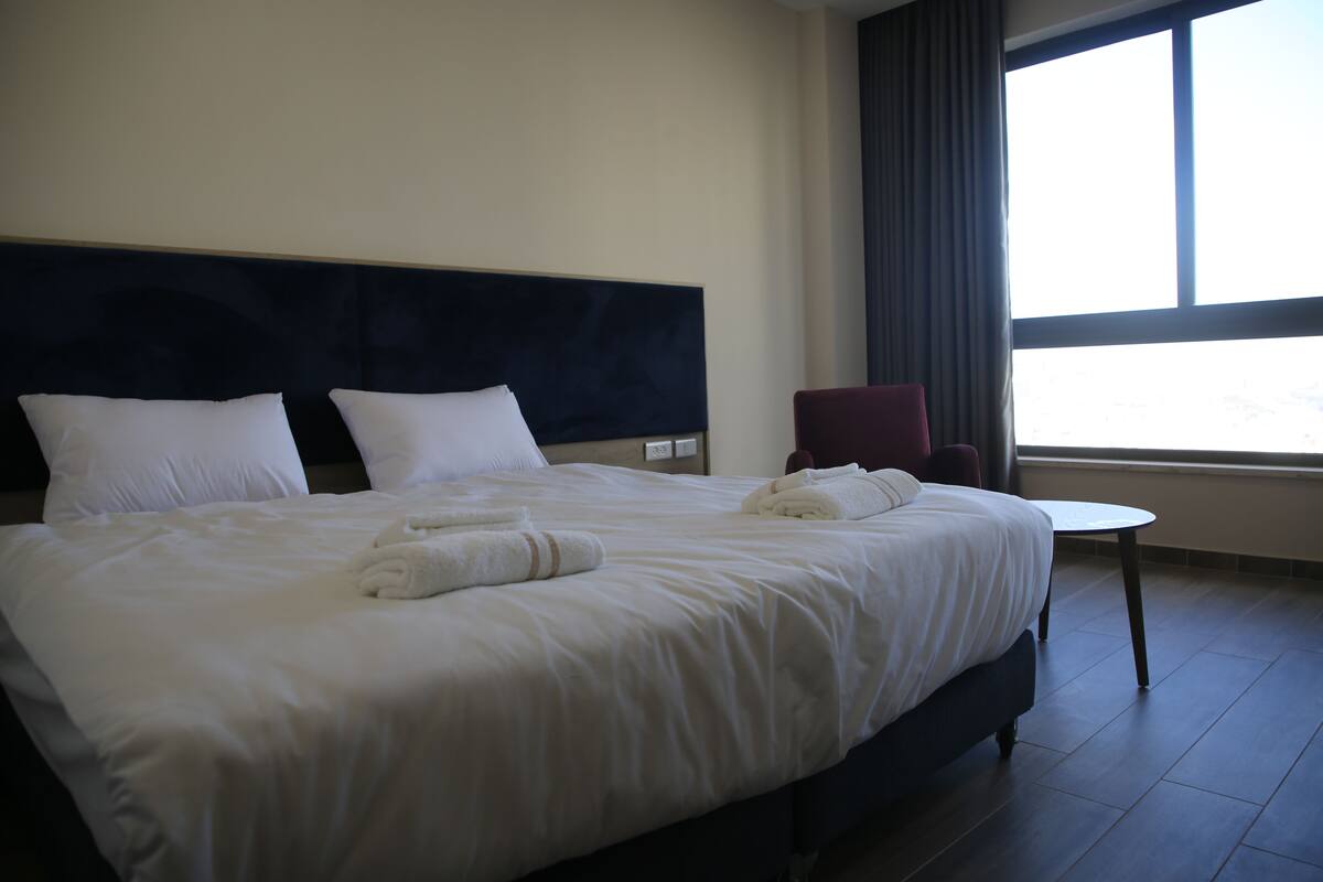 Una cama bien hecha con ropa de cama y toallas blancas, un cabecero oscuro, una mesita redonda pequeña y una silla roja en una habitación de hotel con un ventanal grande.