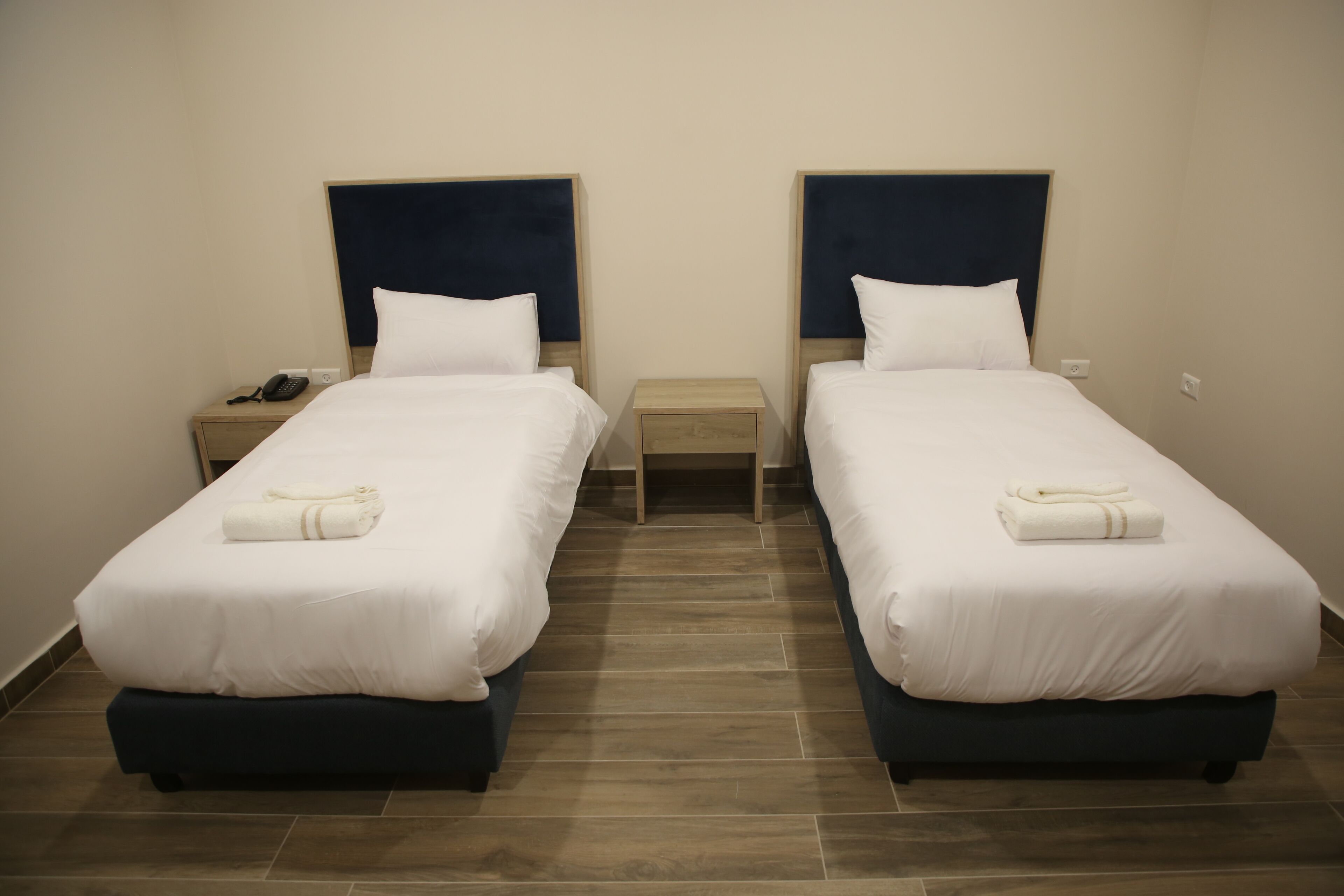 Habitación de hotel con dos camas individuales, cada una con cabecera oscura, ropa de cama blanca y una mesita de noche pequeña con un teléfono y una toalla doblada.