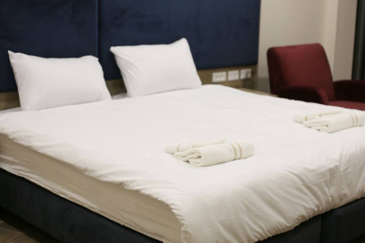 Una cama bien hecha con sábanas y toallas blancas, un cabecero azul oscuro y una silla roja en una habitación de hotel.