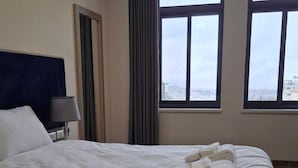 A hotel room with a large bed, a nightstand, a lamp, and a view of the city through large windows. - Saint Francis Hotel (Bethlehem)