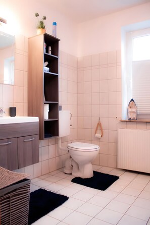 Une salle de bain comprenant des toilettes, une étagère avec des articles de toilette, un radiateur et une fenêtre.