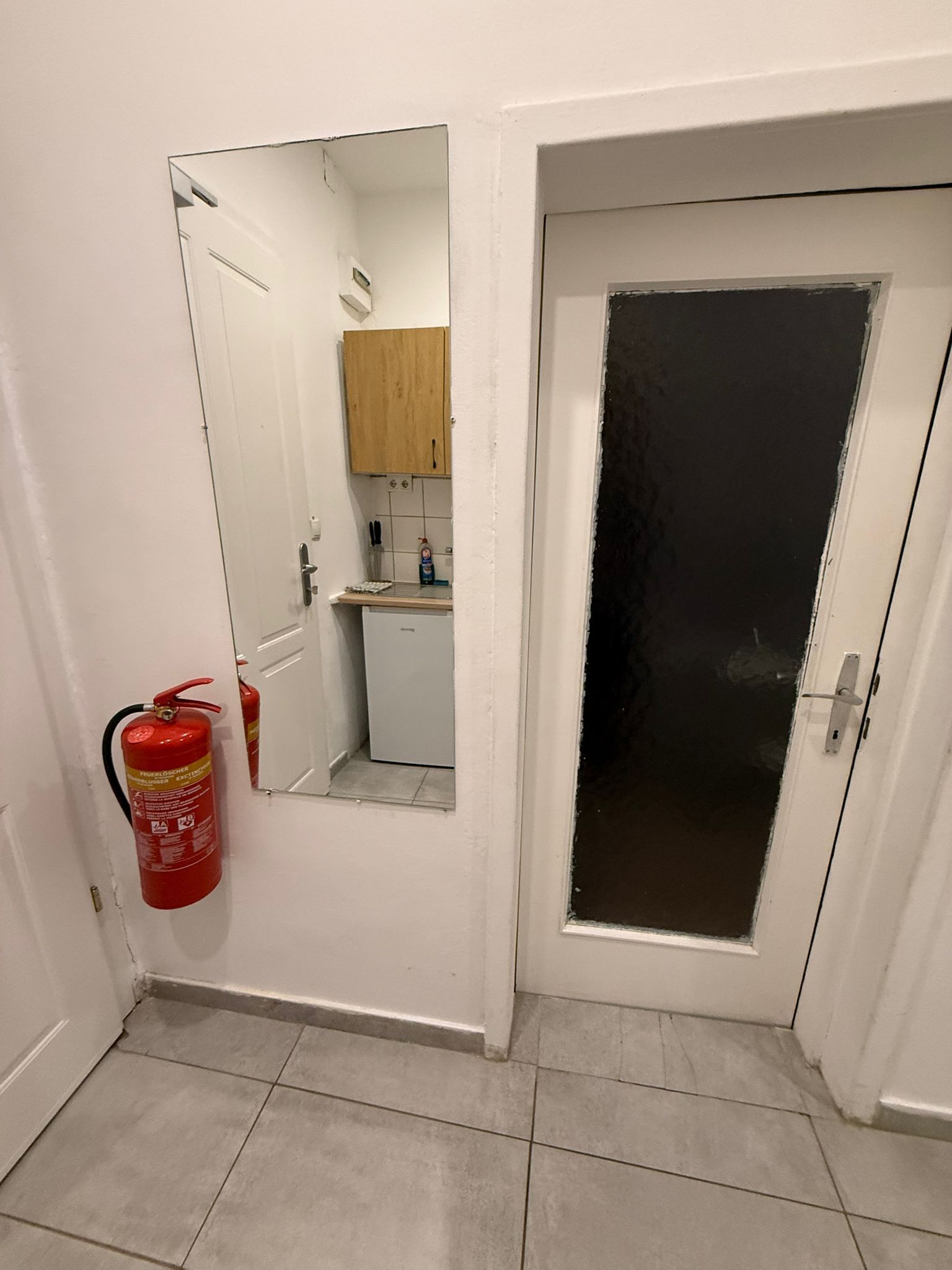 Un pasillo estrecho con una puerta blanca a la derecha, un extintor a la izquierda y una pequeña zona de cocina con un armario de madera y una encimera.