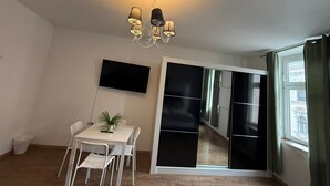 Ein modernes Wohnzimmer mit Essbereich, einem Wand-Fernseher, einer Chandelüstere und einer Glastür zum Balkon.
