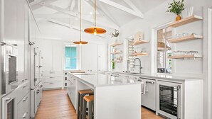 Una cocina moderna con armarios blancos, una isla grande, taburetes de madera, iluminación colgante y estantes empotrados.