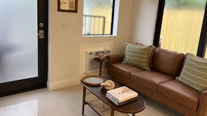 Uma sala de estar com um sofá marrom, uma mesinha, um livro, uma janela e uma porta.