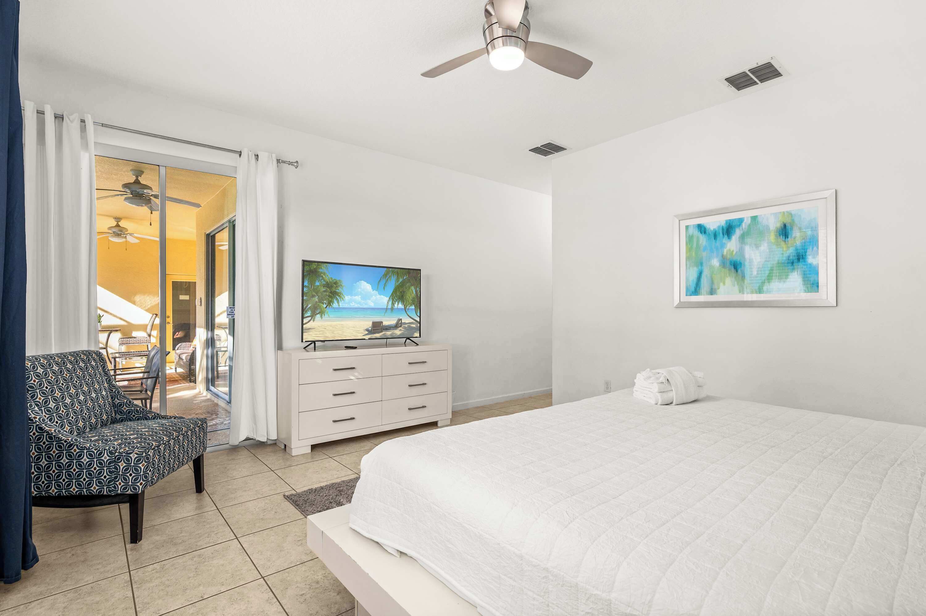A bedroom with a bed, a TV, a ceiling fan, a white dresser, and a painting on the wall.