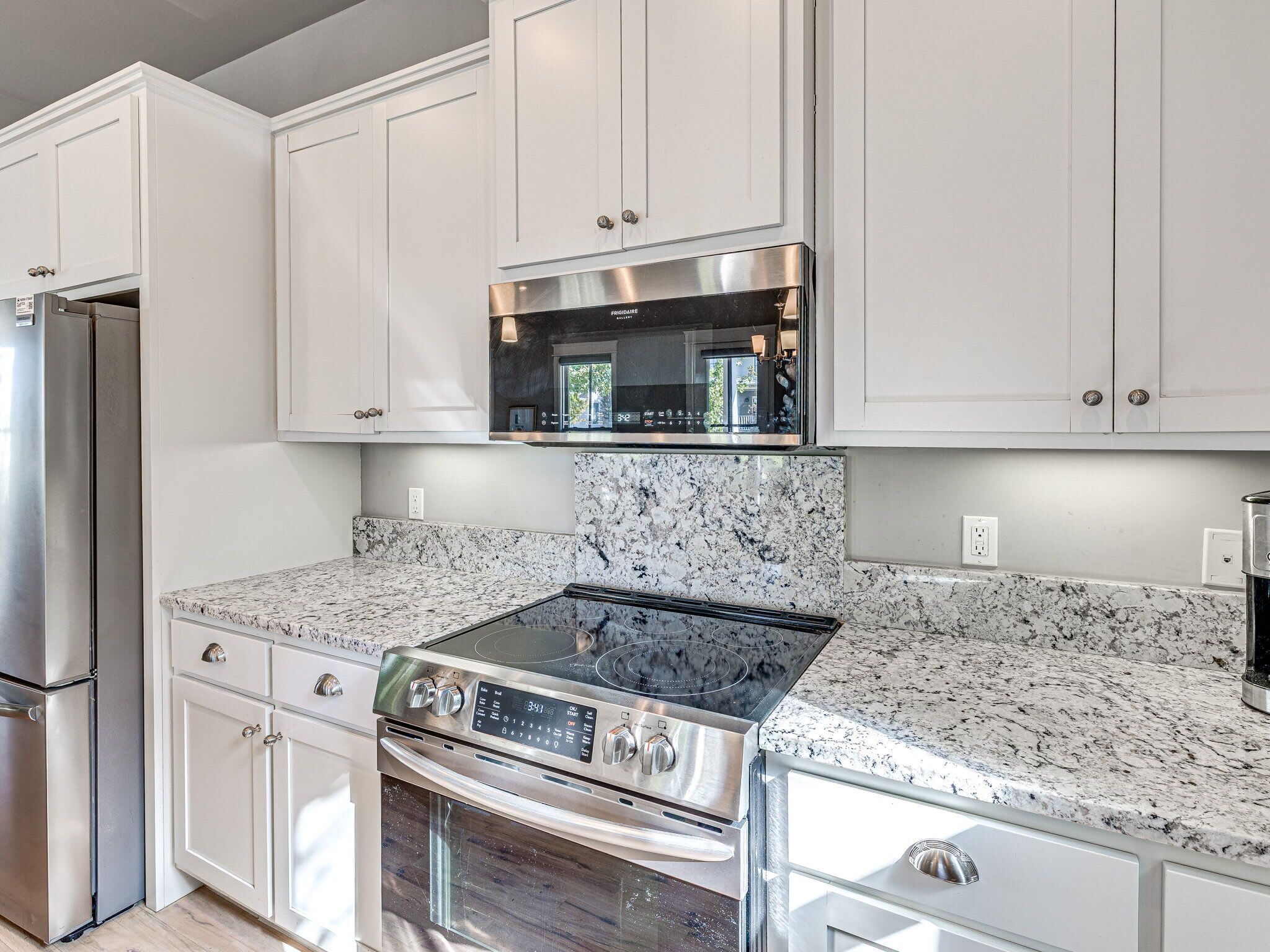 A modern kitchen with white cabinets, a stainless steel refrigerator, a built-in oven, and a granite countertop.