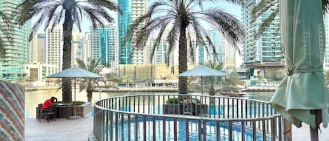 A rooftop pool area with palm trees, a person sitting at a table, and high-rise buildings in the background.