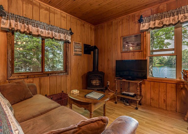 Interior acogedor de una cabaña con paredes de madera, chimenea, televisor y ventanas amplias con vista a un lago.