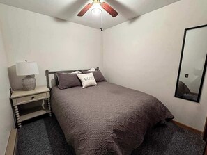 A bedroom with a bed, a nightstand, a lamp, a ceiling fan, and a mirror on the wall.
