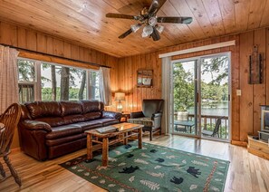 A cozy cabin interior with a leather sofa, a wooden coffee table, and a large window overlooking a lake. - Bouchard Shores Resort - Your Private Northwoods Getaway (Minocqua)