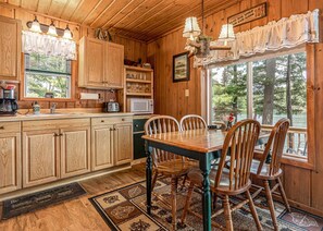 L’intérieur d’une cabane en bois comprend une salle à manger avec une table et des chaises, un coin cuisine avec des placards et des appareils électroménagers, ainsi qu’une grande fenêtre donnant sur une zone boisée.