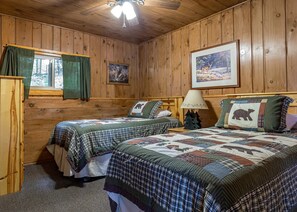 Habitación tipo cabaña con dos camas, techo de madera, ventana con cortinas, un cuadro enmarcado, una lámpara y un aparador.