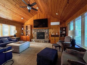 Ein gemütliches Blockhäuschen-Interieur mit Kamin aus Stein, Holzwänden, Deckenventilator und einem großen Fernseher, der darüber angebracht ist.