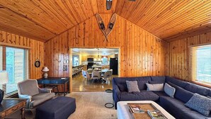 Een gezellig chaletinterieur met een houten plafond, een donkergroene bank, een kleine tafel en een eethoek die door een open deur zichtbaar is.