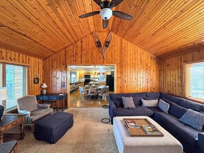 Ein gemütliches Blockhaushaus-Interieur mit Holzbalkendecke, einem dunkelbraunen Sofa, einem kleinen Tisch und einem Essbereich, der durch einen offenen Durchgang sichtbar ist.