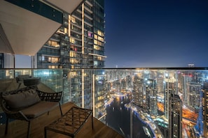 Un immeuble de grande hauteur avec une façade en verre, un balcon doté d’une table et de chaises en bois, et une vue sur le paysage urbain la nuit.