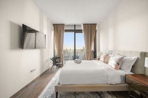 Une chambre d’hôtel moderne avec un grand lit, un téléviseur à écran plat fixé au mur, une vue sur la ville par la fenêtre et une petite table avec une lampe.