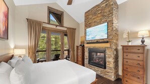 A bedroom with a large bed, a stone fireplace, a TV mounted on the wall, and a view of the outdoors. - Charming Ski Condo Near Keystone with Fireplace & Scenic Balcony (Keystone)