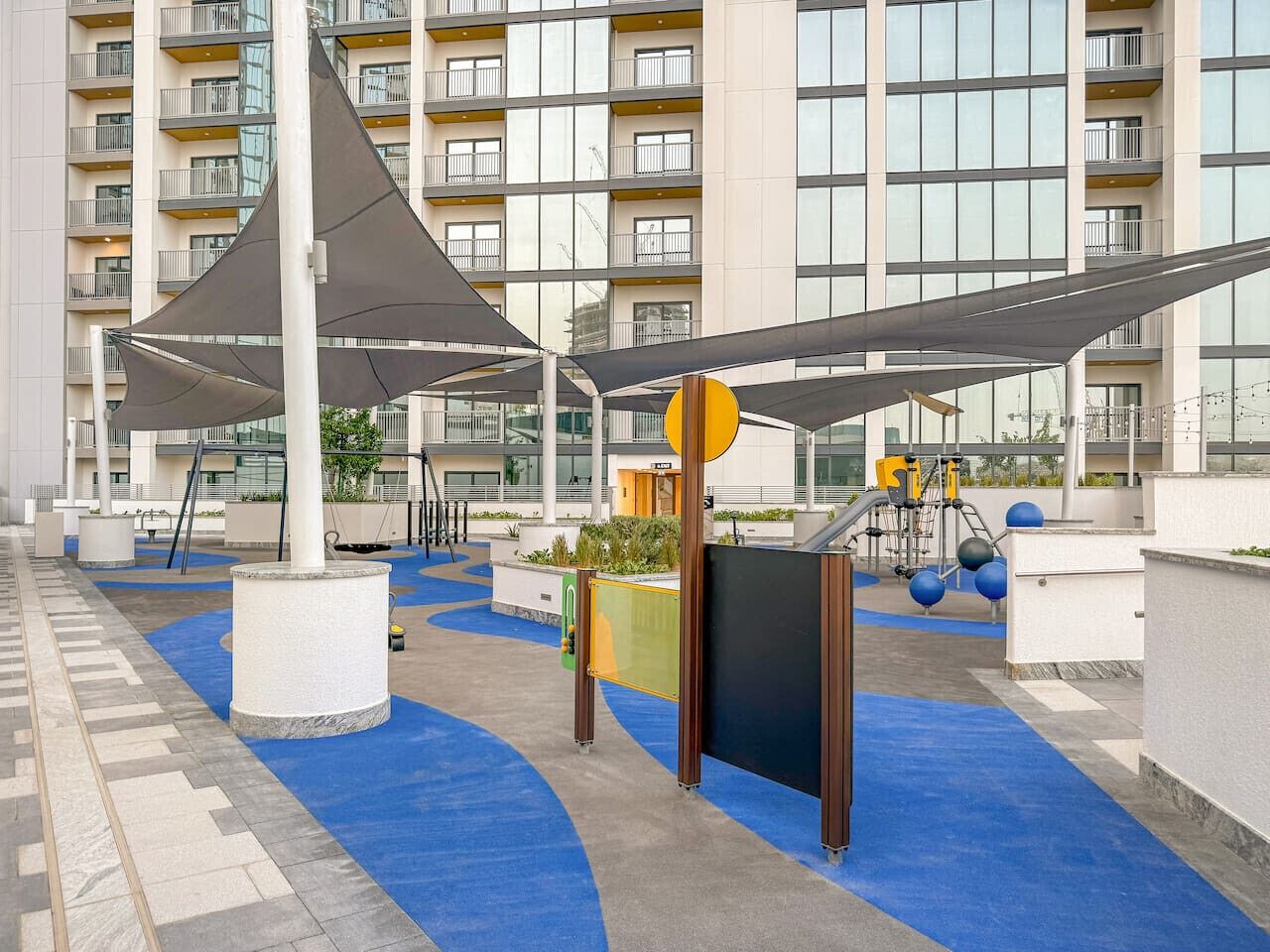 Zona de juegos con piso de caucho azul, una gran vela de sombra gris y un edificio con balcones al fondo.