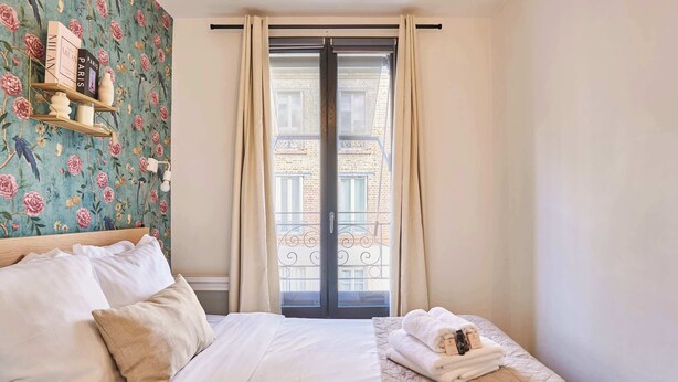 A hotel room with a bed, a small table with decorative items, a window with curtains, and a view of the outside balcony.