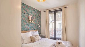 A neatly made bed with a floral wallpaper, a shelf with toiletries, and a view of a balcony.