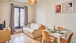 A modern living room with a sofa, dining table set for four, and a large window with curtains.
