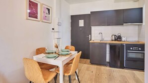 A compact kitchen with modern appliances, a dining area with a white table and brown chairs, and two framed artworks on the wall.