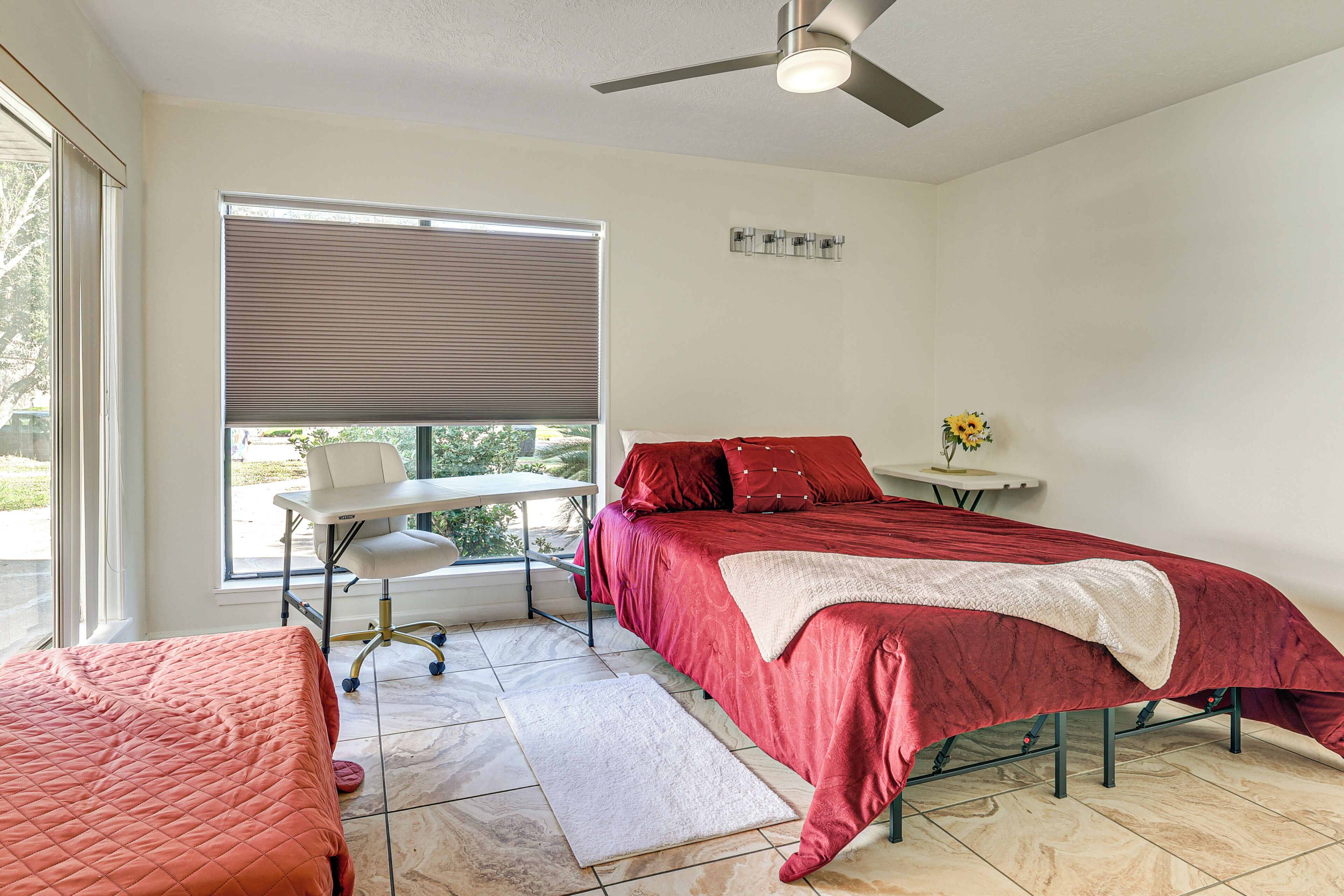 A bedroom with a bed, a desk, a chair, a window with blinds, and a ceiling fan.