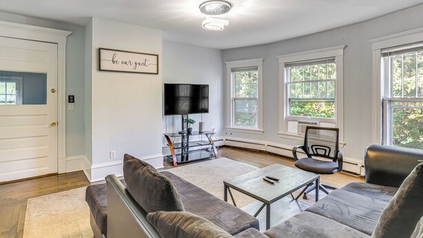 A modern living room with a gray sectional sofa, a coffee table, and a TV. There are large windows with a view of trees. - 2 Mi to Dtwn! Walkable Minneapolis Apt w/ Deck (Minneapolis)