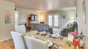 A dining area with a wooden table set for four, surrounded by white chairs. There is a gray sofa in the background, a television on a stand, and a door leading to another room. - 5 Mi to Dtwn: Tucson Townhome w/ Pool Access! (Tucson)