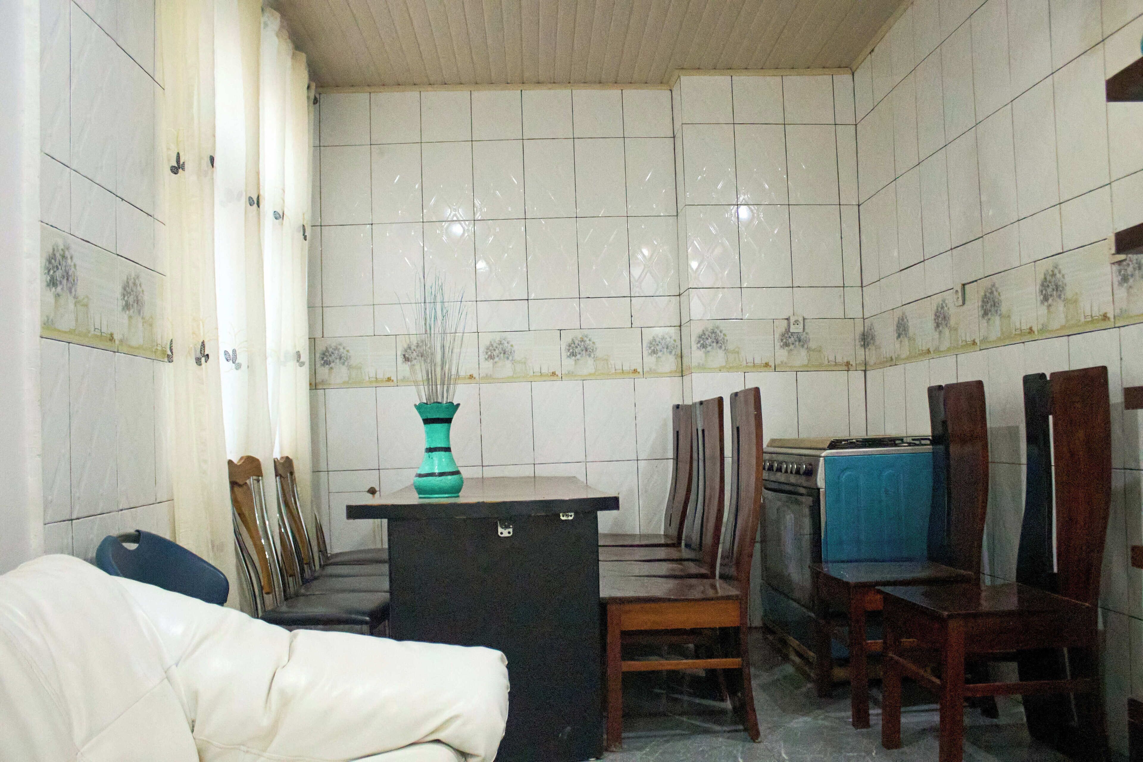 A compact kitchen with white tiled walls, a central island with a teal vase, wooden chairs, and a blue refrigerator.